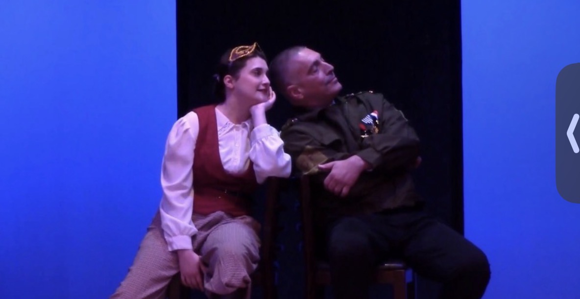 Crowned figure leaning on a military officer under blue stage light