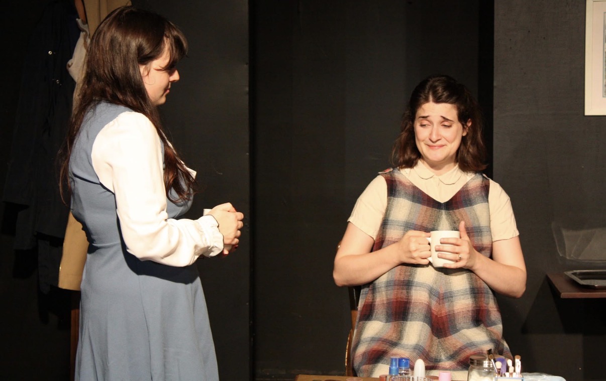Two women in 1960s dress, one holding a mug in an emotional moment